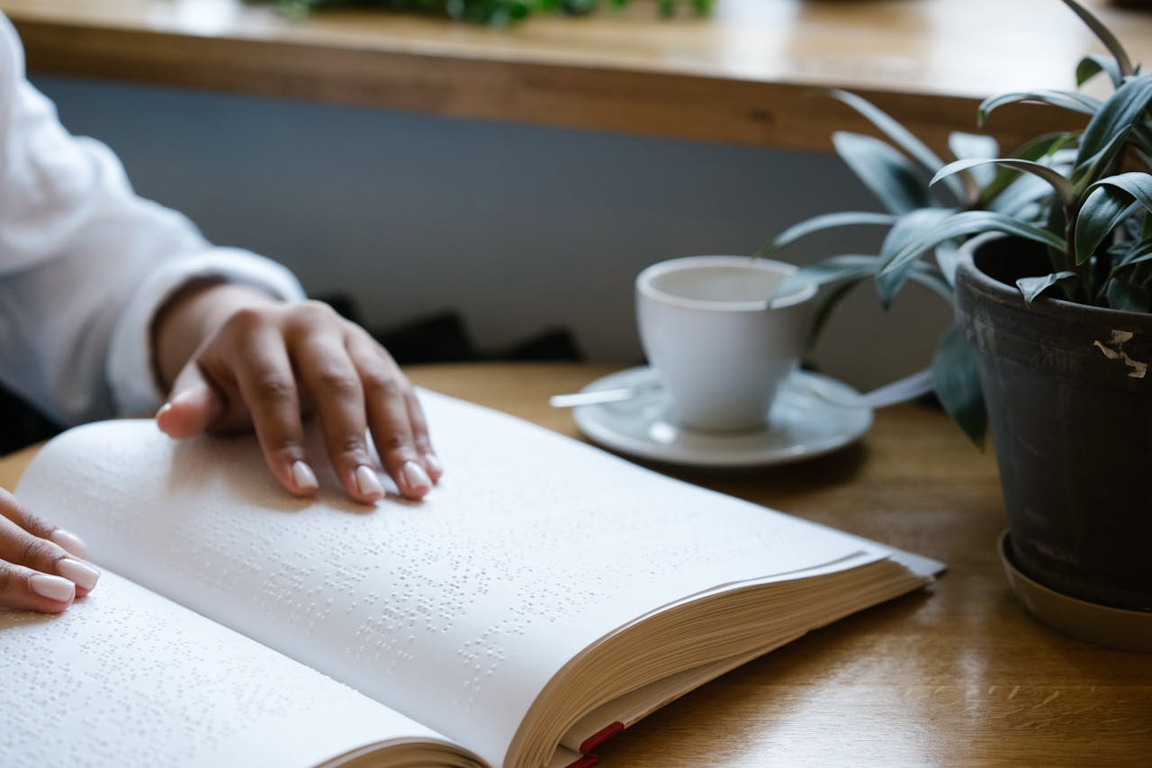 A person reads a Braille book at a wooden table next to a plant and coffee.